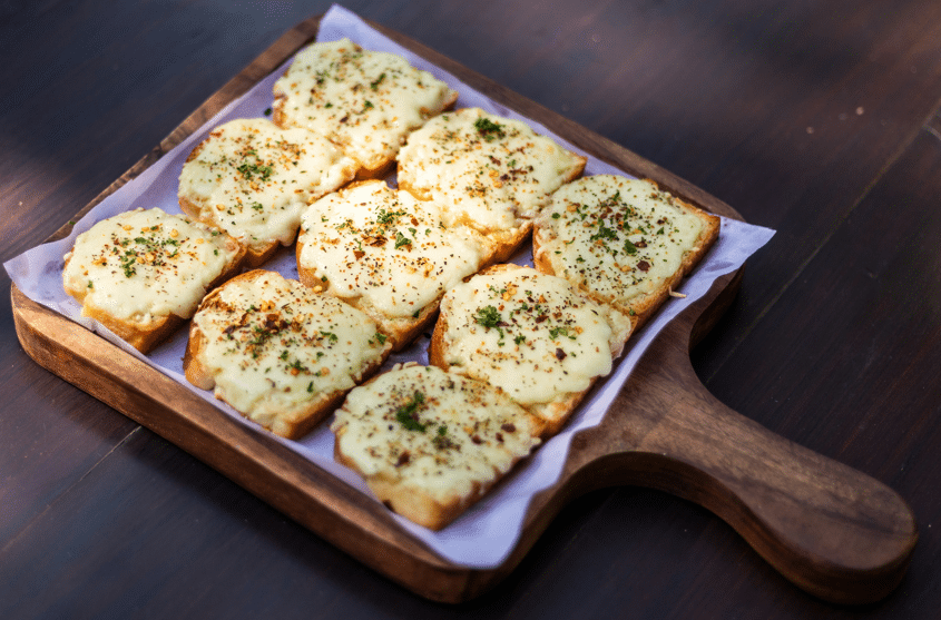 Golden-brown garlic bread topped with melted cheese and sprinkled with herbs, served on a wooden tray